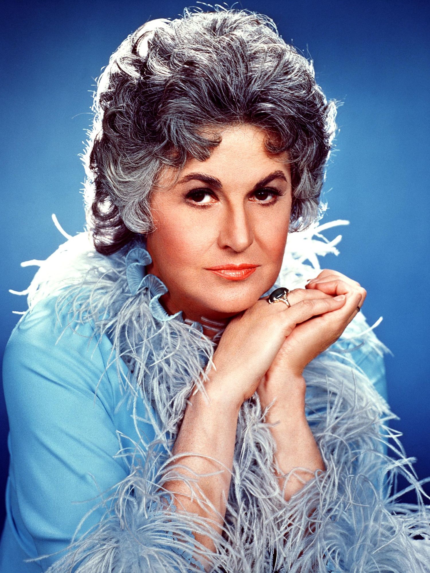 Actress Bea Arthur poses in a light blue dressing gown with a feathered collar and cuffs against a darker blue backdrop.