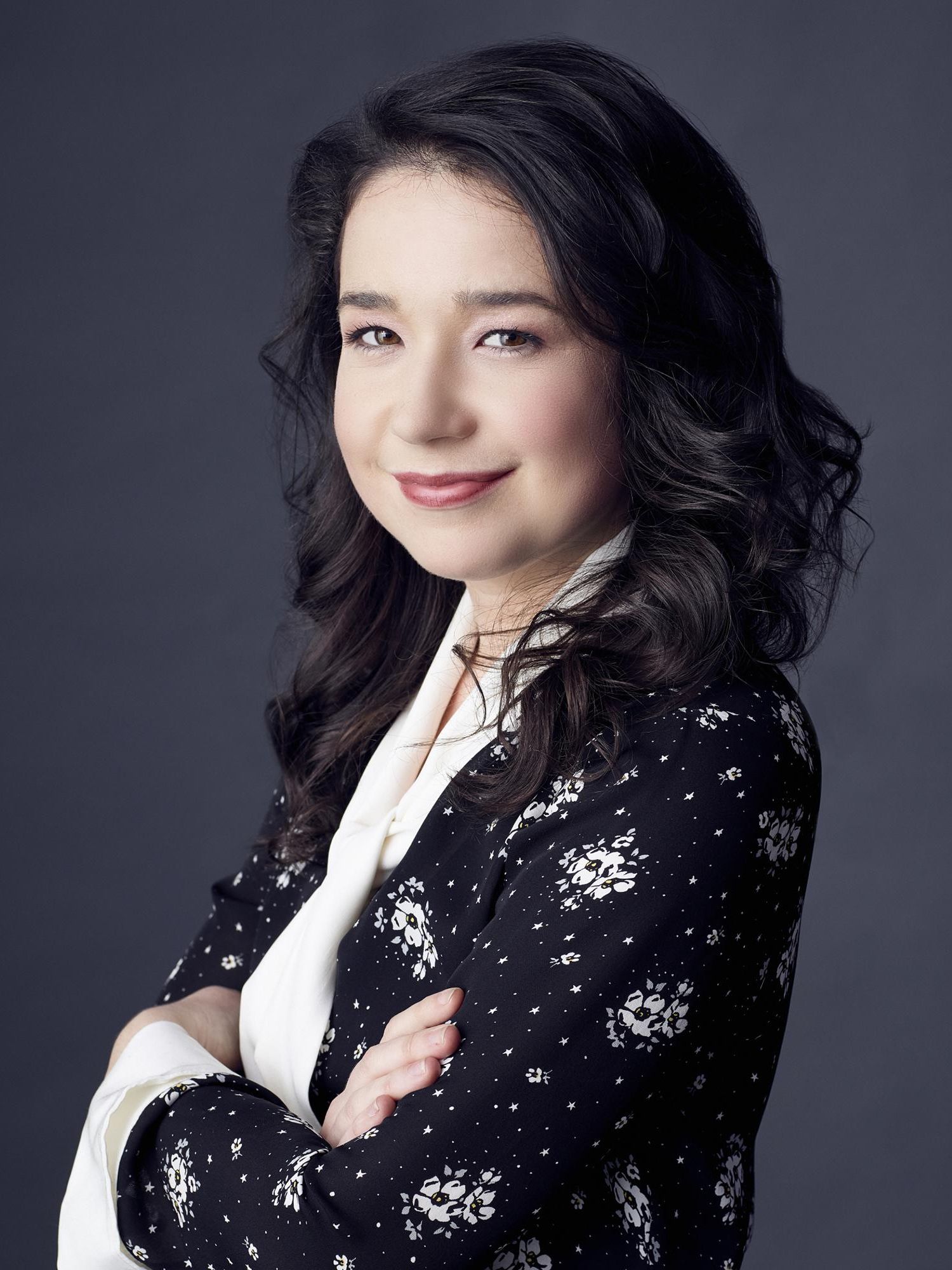 Sarah Steele of The Good Fight poses against a gray backdrop wearing a white blouse and flower patterned navy blazer.