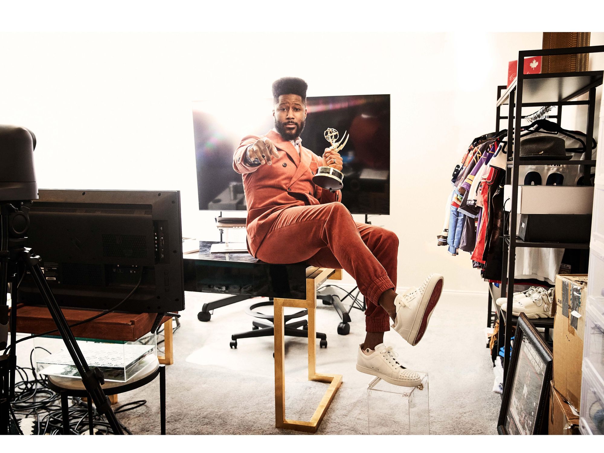Former NFL player Nate Burleson wearing an orange suit and holding an Emmy in his office, with sports jerseys on a rack to his left and camera equipment to his right.