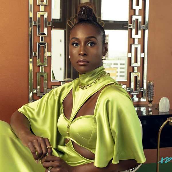 A Black woman, Issa Rae, sits in a salon chair in a regal, lime green Prabal Gurung gown with cut outs on both sides of her chest and rib cage. The flowing short sleeves stop at her elbows. She delicately holds her fingers in her lap and stares directly into the camera. She's sitting in front of a mirror, with hair brushes behind her on the counter and a hot press stove