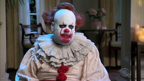 A recognizable CBS star dressed as Pennywise with white face paint, a red clown wig, and a white clown suit with ruffles around the neck.