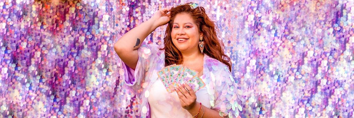 astrologer La Bruja del 305 stands with tarot cards fanned in her hand in front of purple toned large sequined wall 