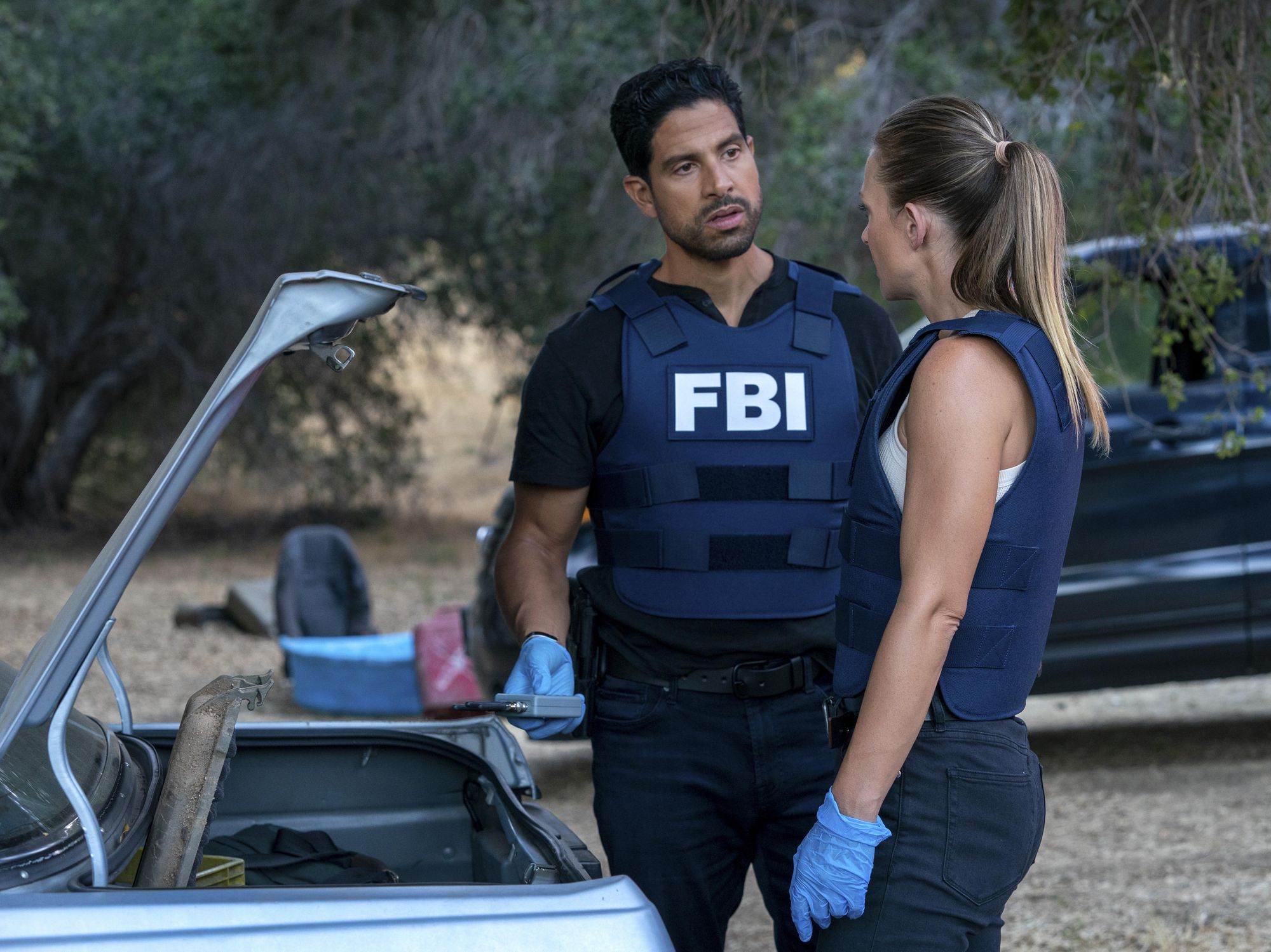 Adam Rodriguez as Luke Alvez and AJ Cook as Jennifer JJ Jareau  stand next to the open trunk of a car wearing their FBI vests and having a serious conversation.
