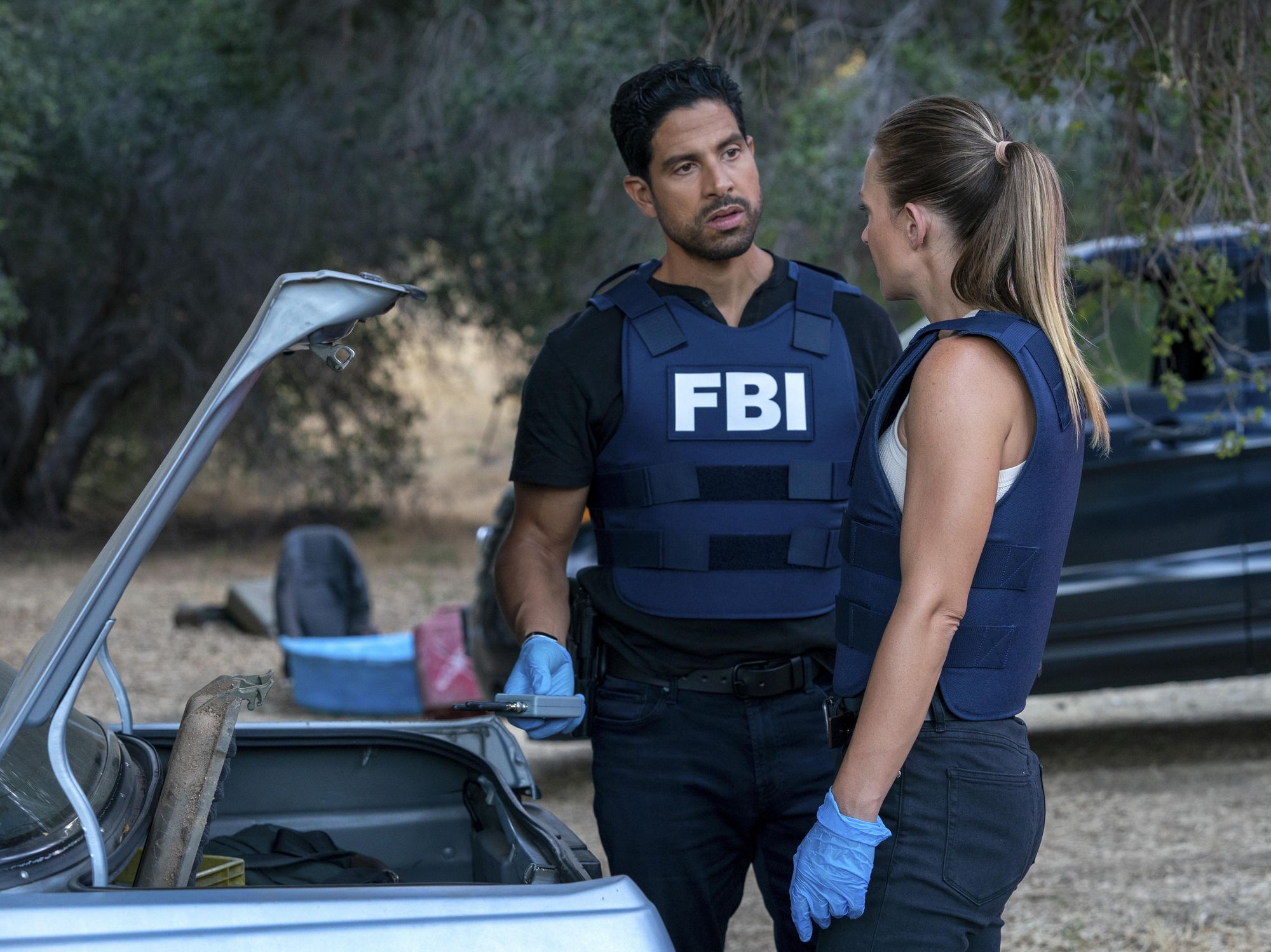 Adam Rodriguez as Luke Alvez and AJ Cook as Jennifer JJ Jareau stand next to the open trunk of a car wearing their FBI vests and having a serious conversation.