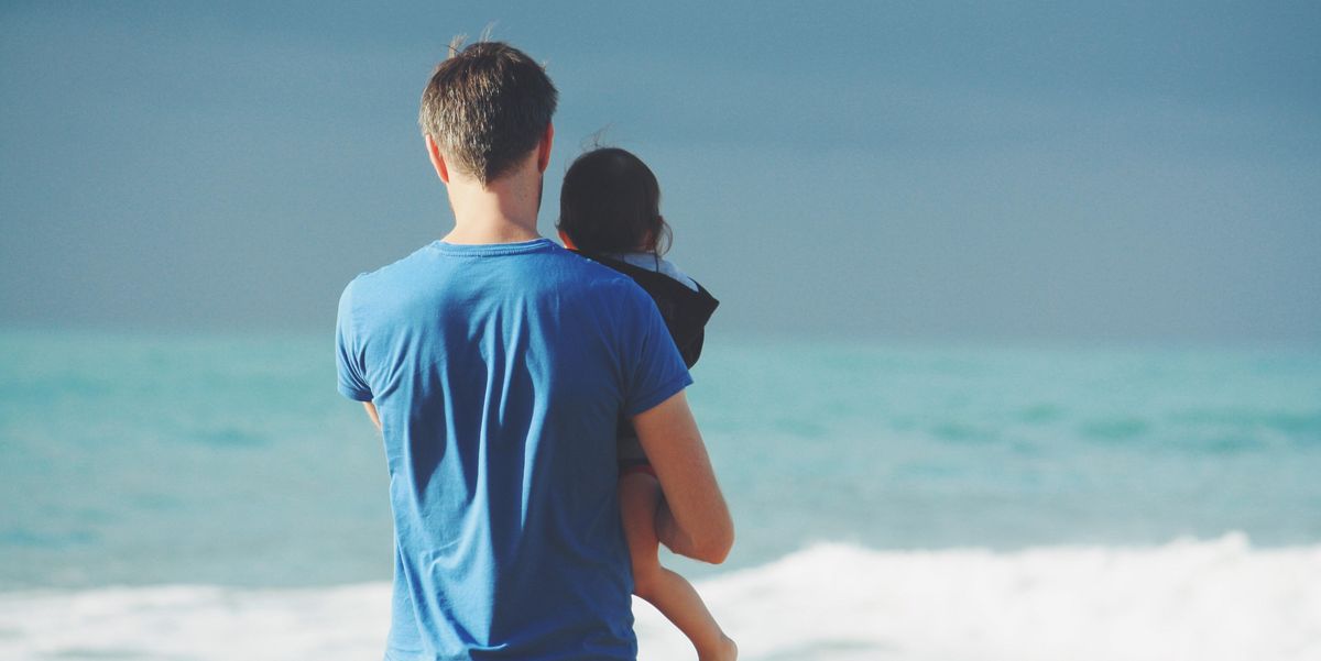 man in blue t-shirt holding toddler in black hooded jacket near ocean under blue sky