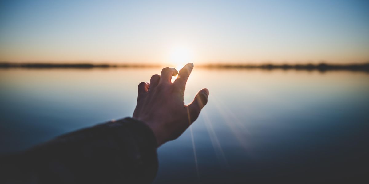 man's hand reaching toward the setting sun over a lake