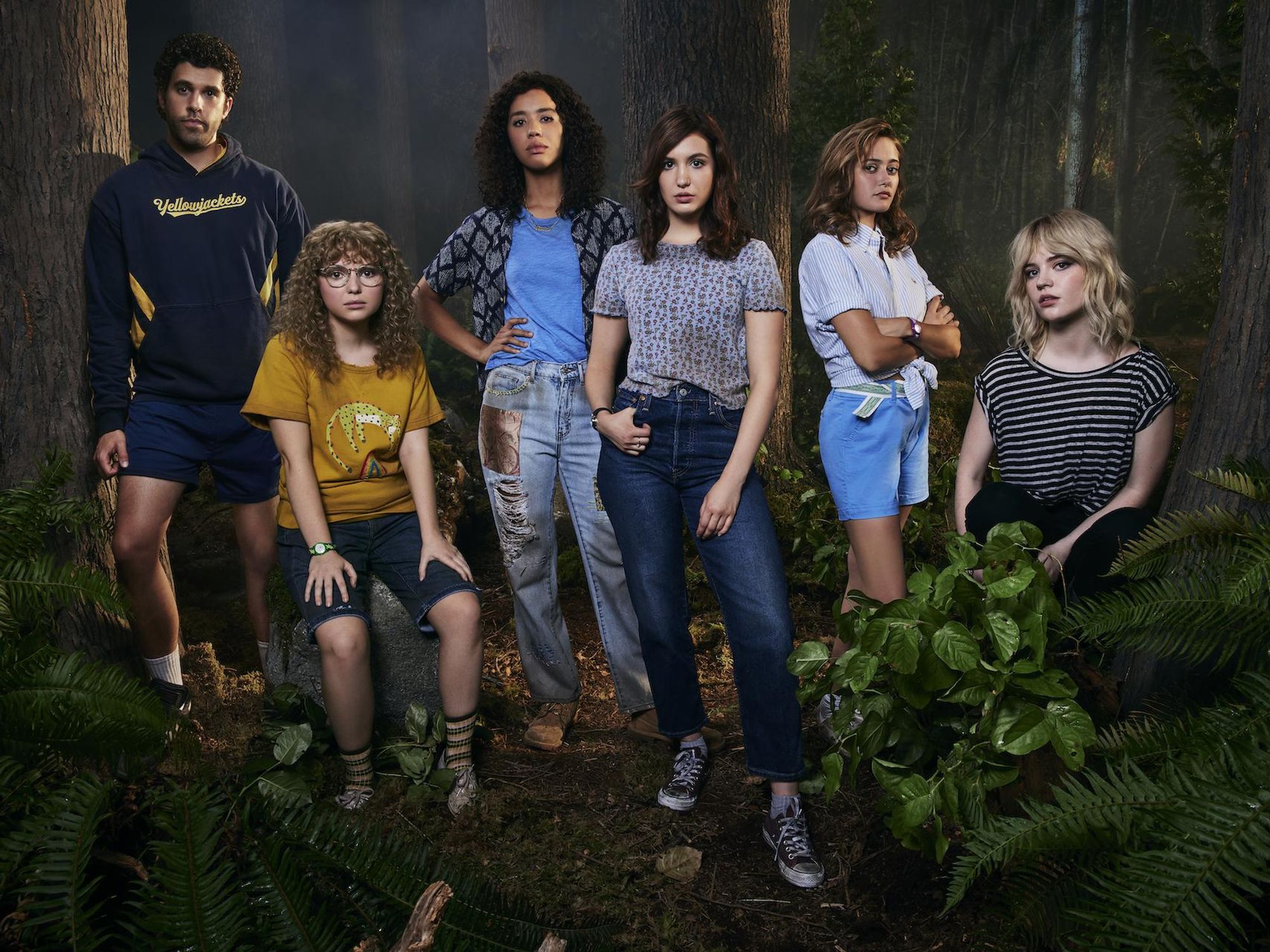 A group of teenagers stand in a wooded area surrounded by plants and trees looking serious.