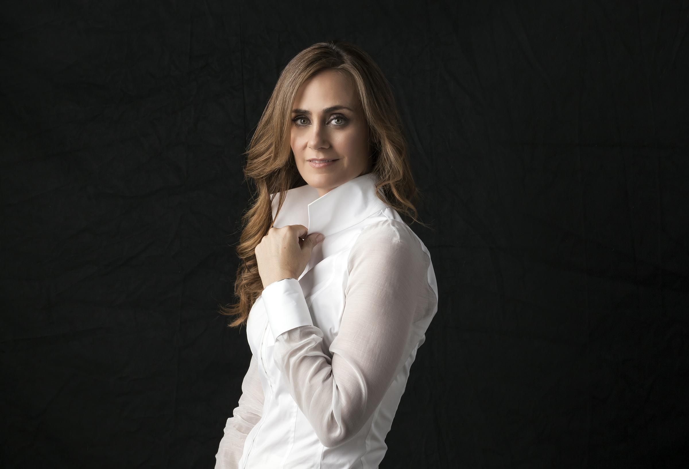 Actress Diane Farr of the new CBS series Fire Country wears a high collared white shirt against a charcoal backdrop.