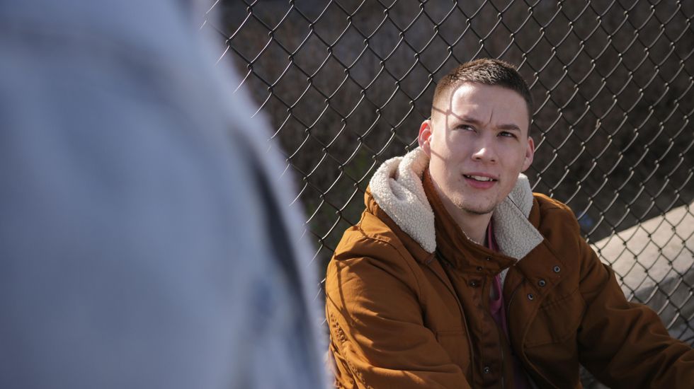 Young man wearing brown jacket sitting near gray link fence