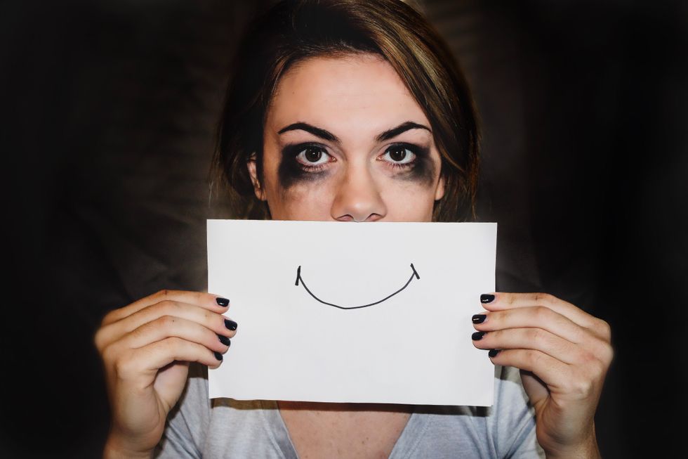 person with smudged eyeliner holding white printer paper over their mouth with a drawn smile