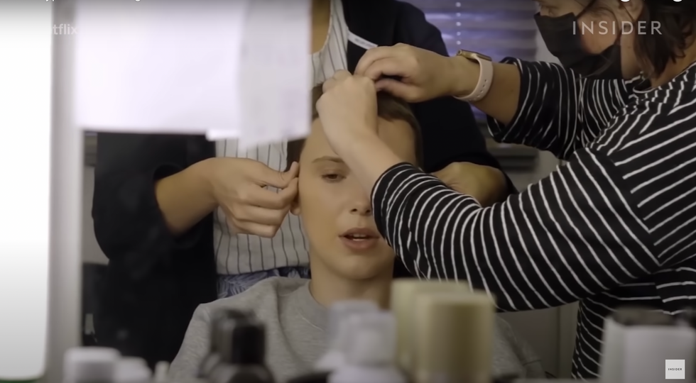 Actress millie bobby brown seated and having a wig applied by two wigmakers.