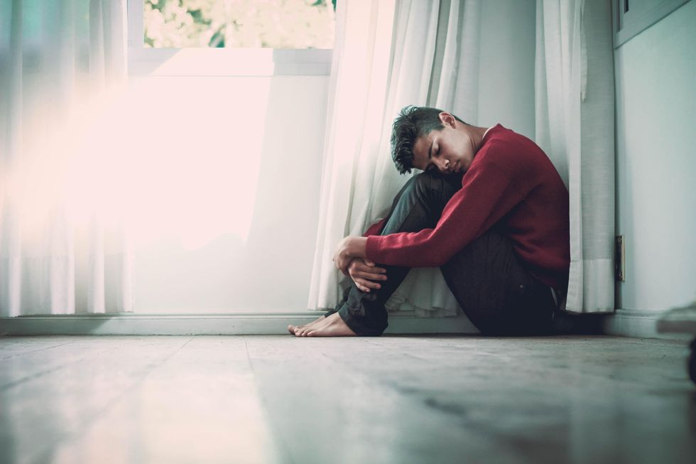 A young man crouched on the floor by a window, eyes closed, contemplating his mental health.