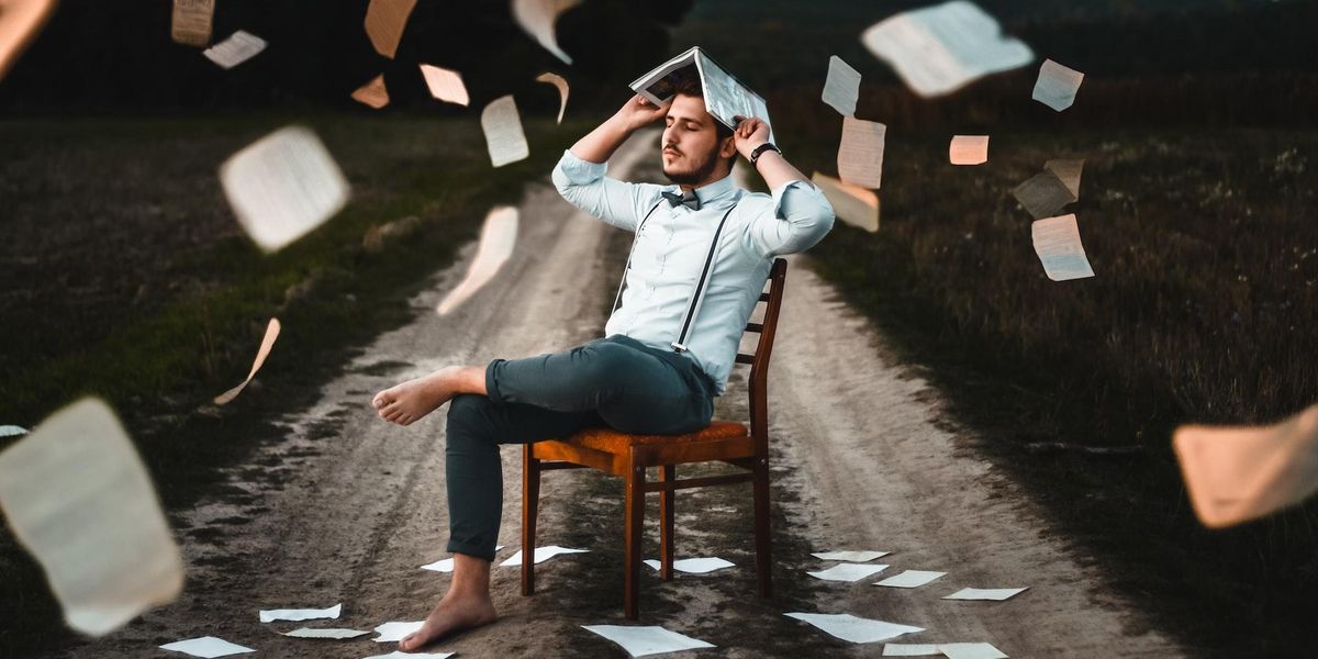 Man sits in a chair barefoot, eyes closed on a dirt road as pages of books fly around him