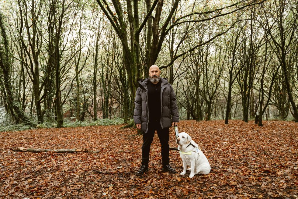 The Blind Poet Dave Steel standing in winter clothes in a copse in fall with his seeing eye labrador retriever sitting by his side.