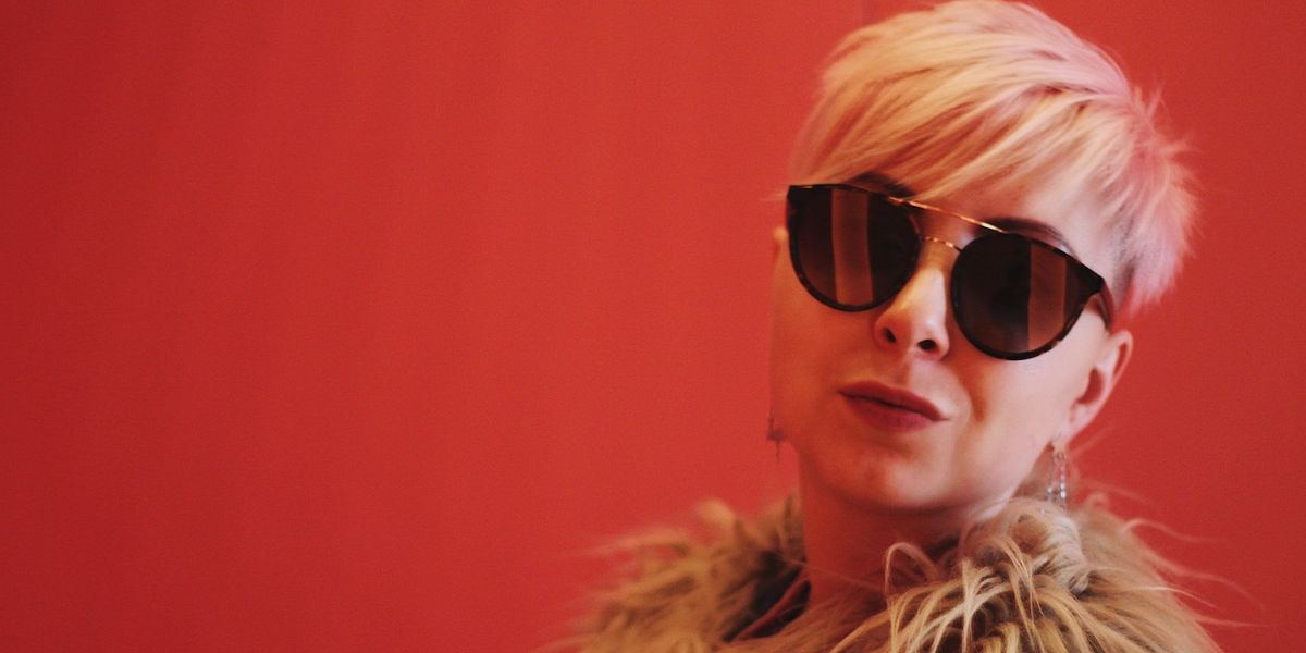 A blonde woman with sunglasses poses into the camera against a red background