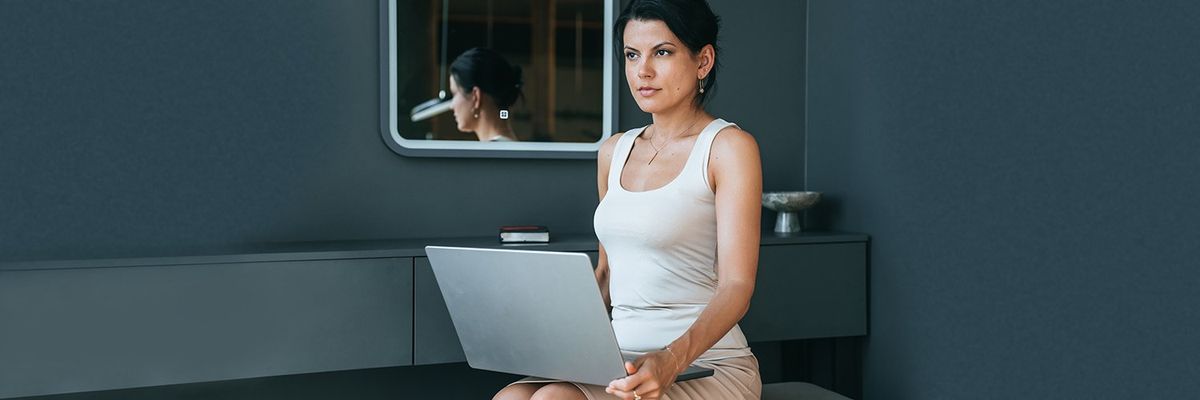 woman with dark hair wearing a business skirt and tank top sitting with an open laptop on her lap 