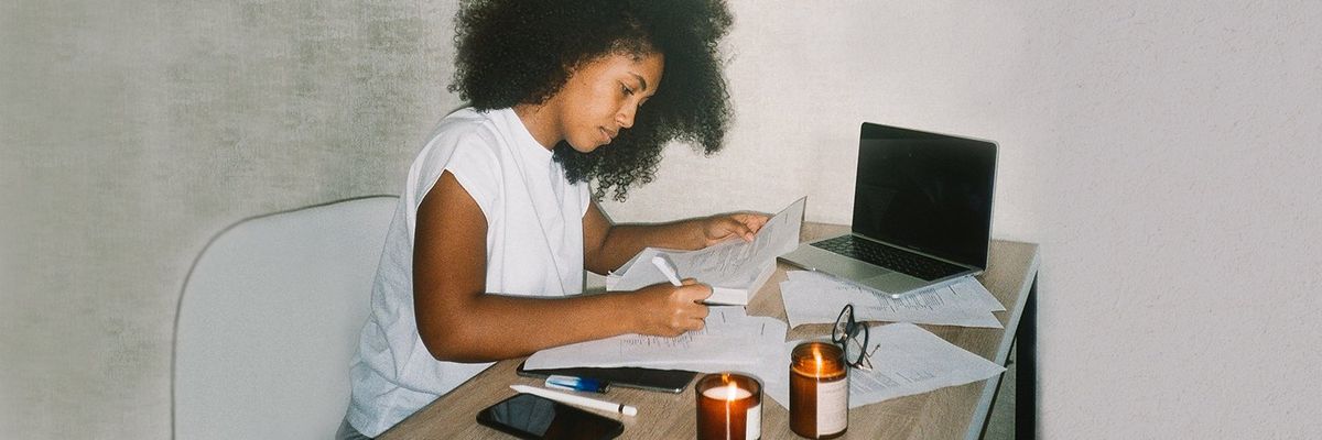 a curly haired woman at a desk writing on a piece of paper with a laptop, a smartphone and a candle beside her