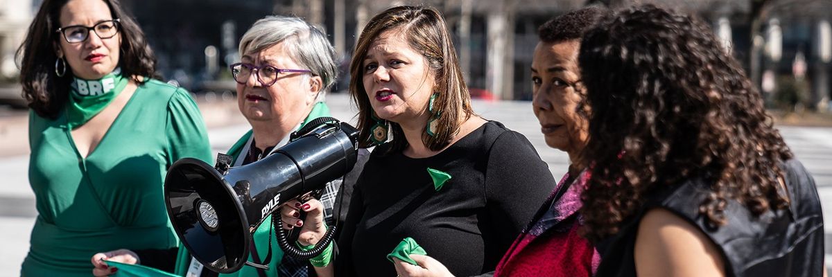 five women wearing green clothing and accessories are gathered with one speaking into a bullhorn 