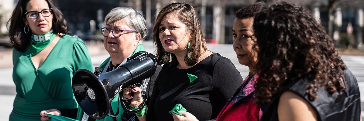 five women wearing green clothing and accessories are gathered with one speaking into a bullhorn