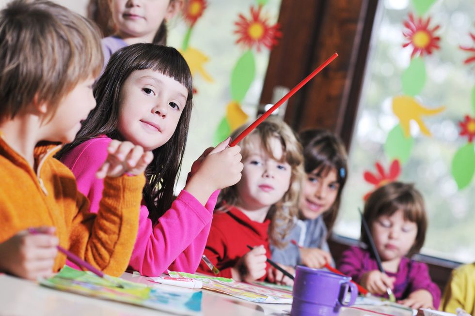 In a group of 5 young children sitting around a table painting, one girl in a pink shirt with brown hair and brown eyes looks at the camera with a special look in her eye.