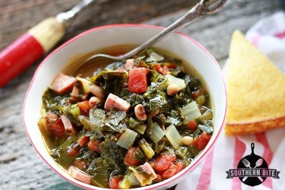 A white bowl of Potlikker Soup with collards, ham, tomatoes, black-eyed peas and cornbread on the side.