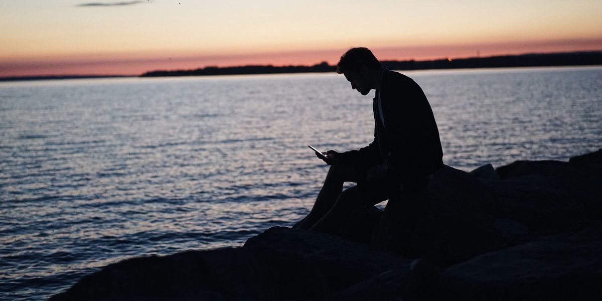 Silhouette of a man sitting by the ocean looking at his phone