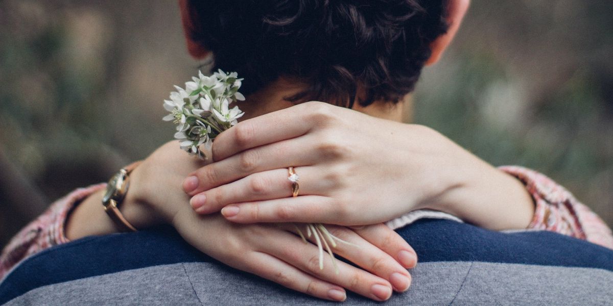 A woman has her hands around a man's neck. She wears an engagement ring and holds a small bouquet of white flowers. 