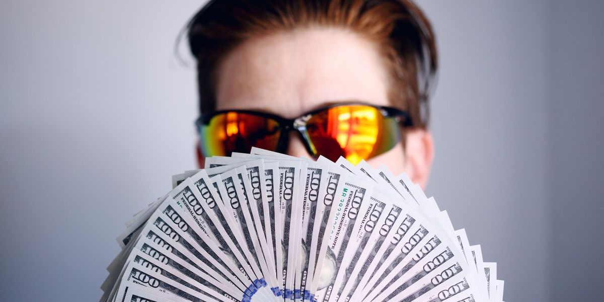 A young man with orange tinted sunglasses fans out a large amount of one hundred dollar bills