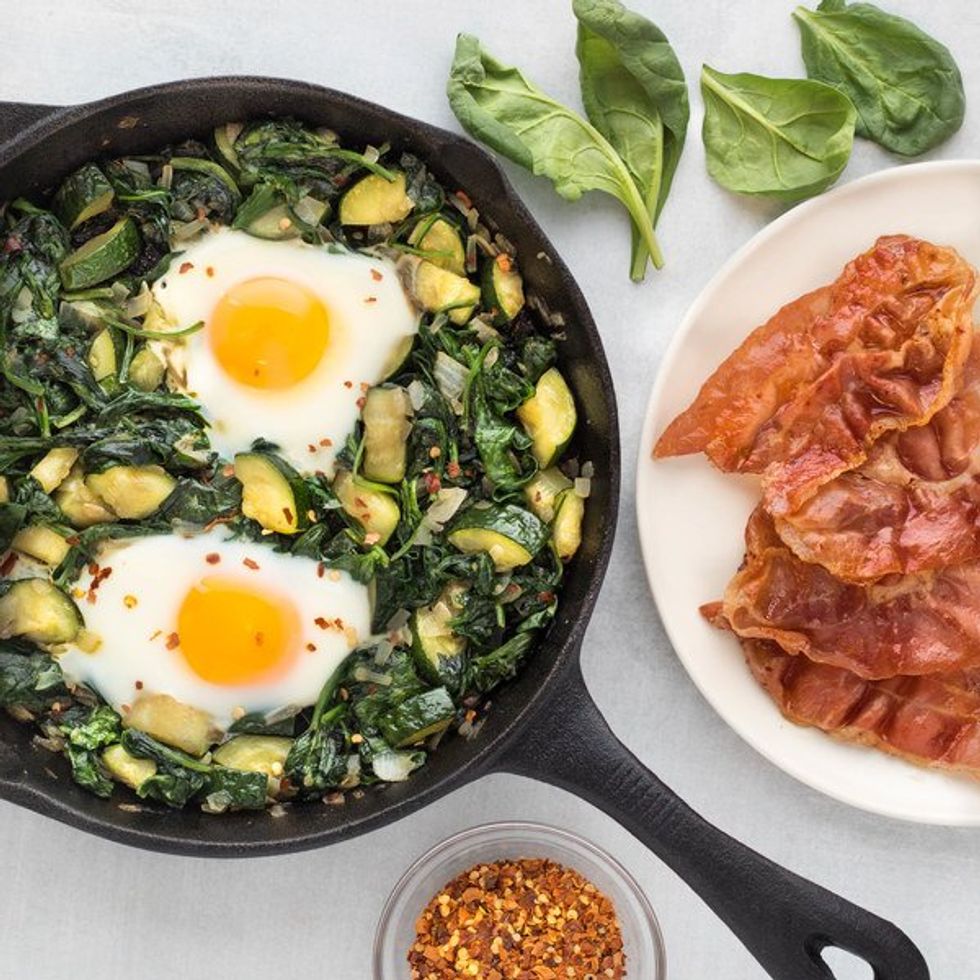 eggs and green veggies in a skillet, plate of bacon