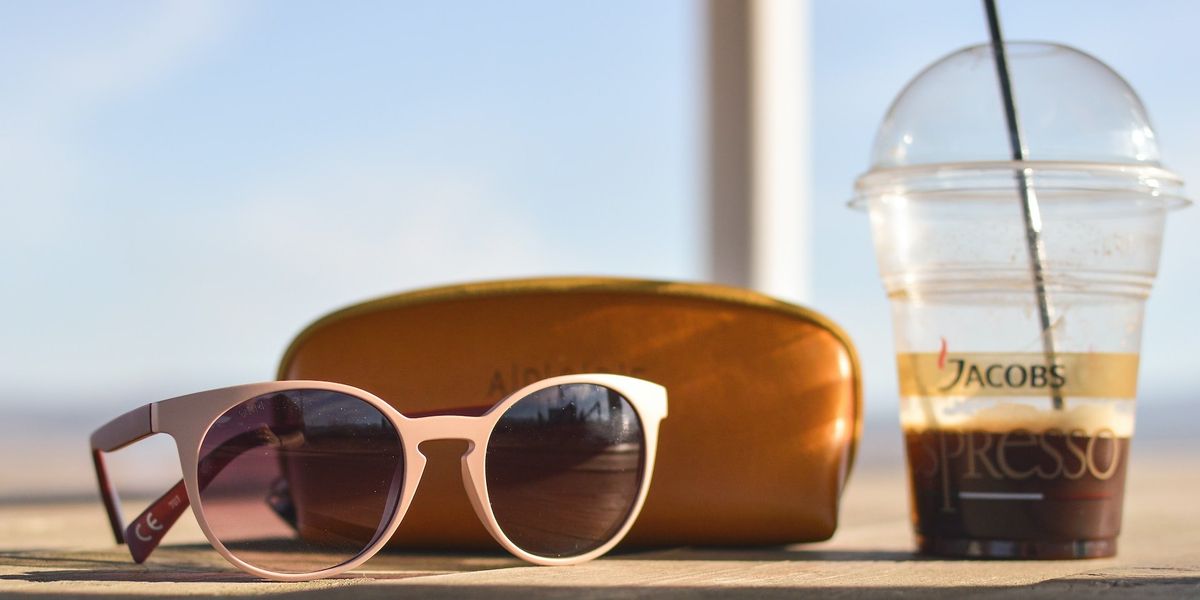 A pair of sunglasses, their case and an iced espresso coffee are placed on top of a counter