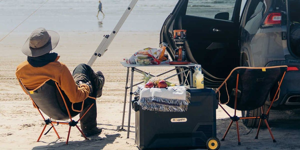 a photo of a man sitting on the beach fishing with a portable refrigerator next to him