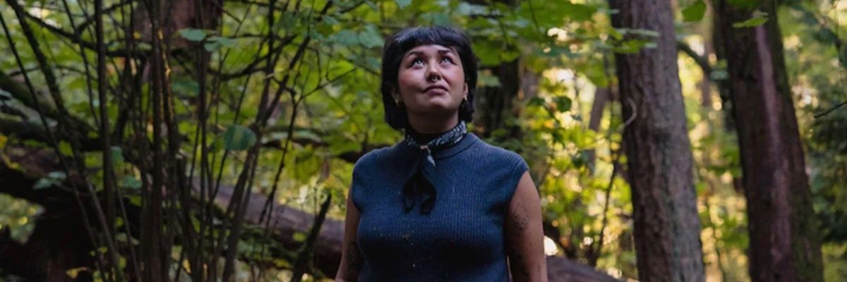 woman with dark short hair in dark blue sleeveless top stands in the middle of a forest looking up at the sky