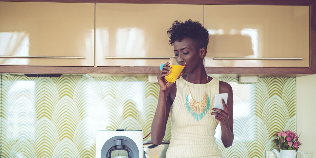 Black-woman-sitting-on-her-kitchen-counter-drinking-juice-while-holding-her-phone
