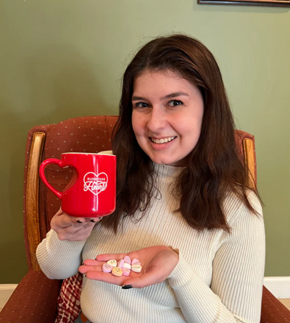 Woman sits holding a handful of heart candies and a red coffee mug that says Bless Your Heart.