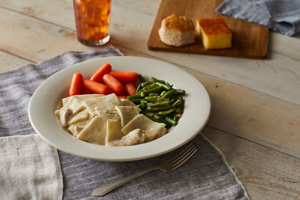 A white plate with chicken and dumplins, green beans and carrots sits on a table besides a biscuit and piece of cornbread.
