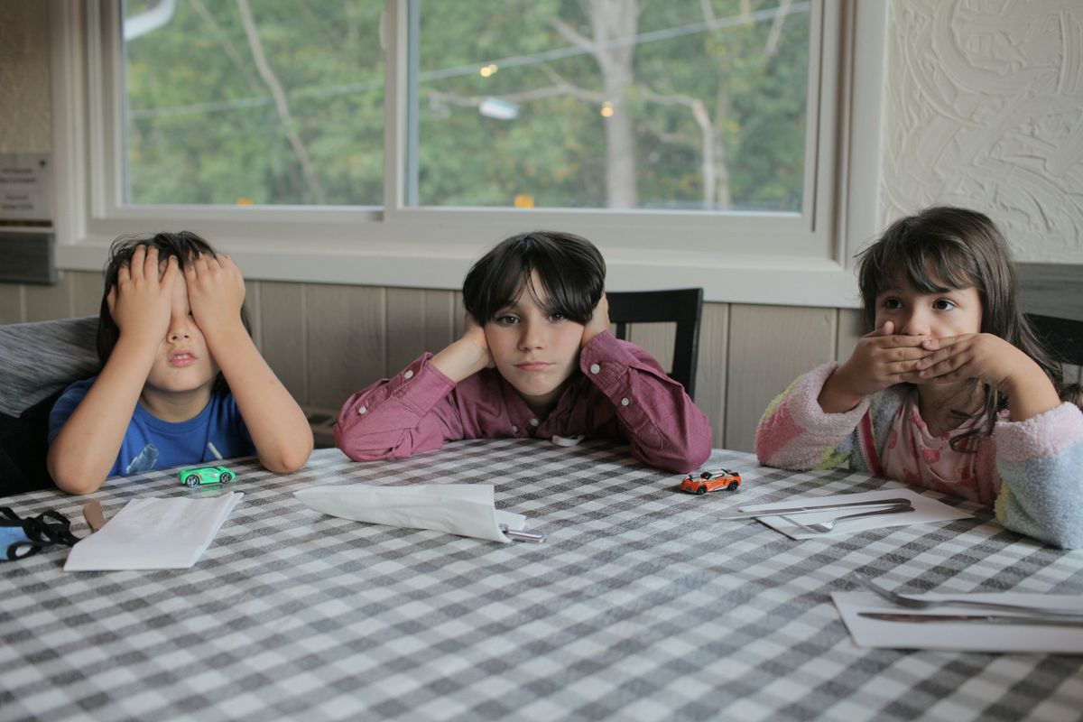 three kids at a table, one with hands over eyes, one with hands over ears, one with hands over mouth