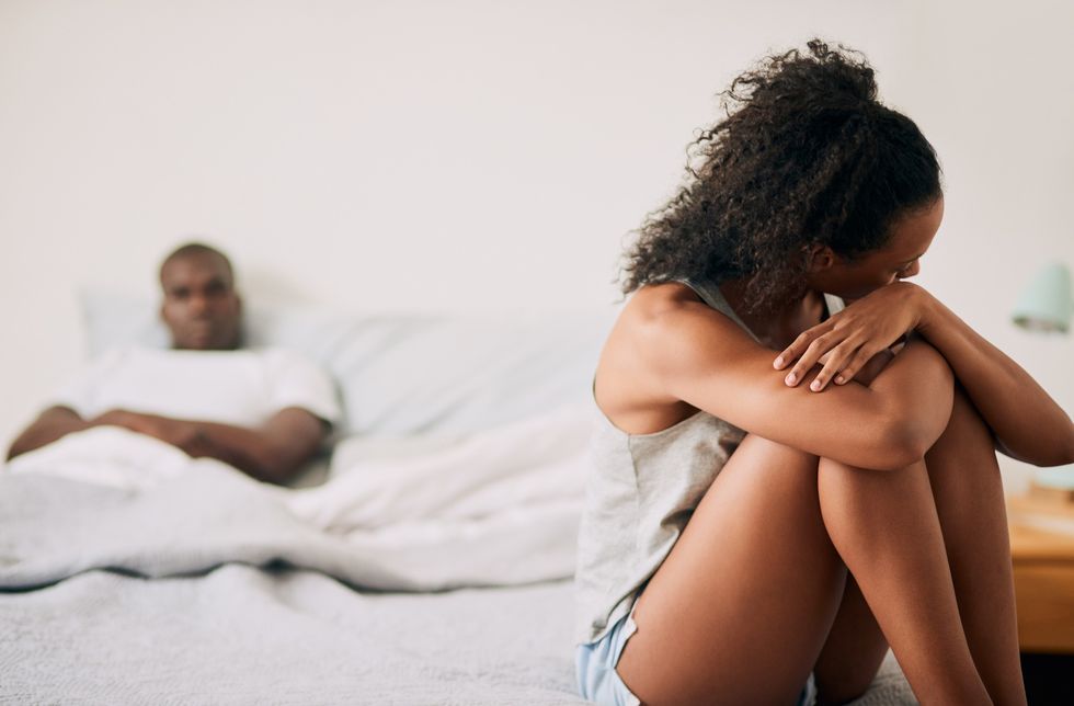 Woman-upset-sitting-on-the-edge-of-the-bed-with-her-partner-in-the-background