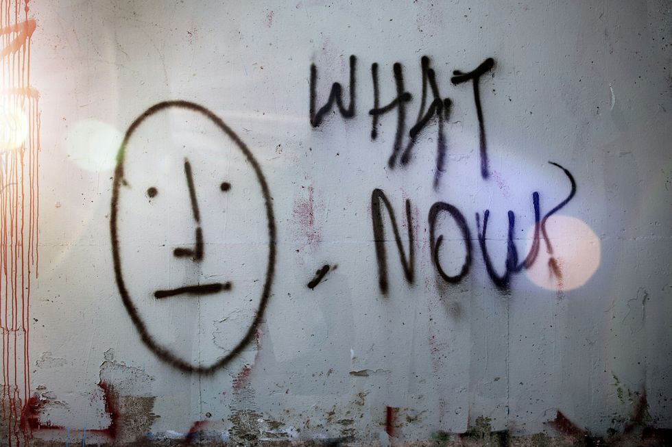 a stick figure face and the words "What Now" spray painted in black paint on a w