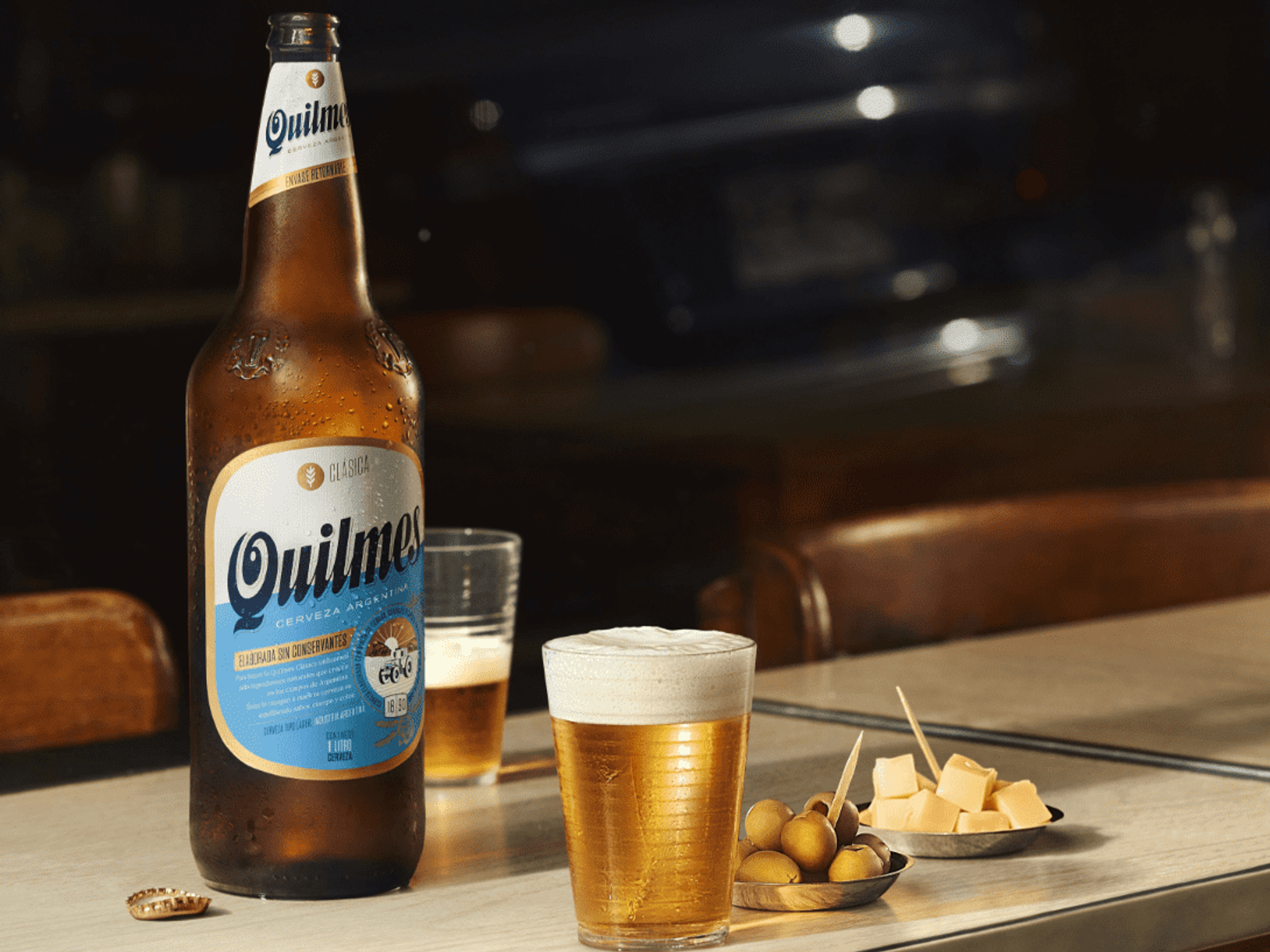 Argentine 'Quilmes' beer on a bar table, with glasses, olives, and cheeses.