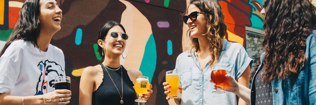 Four Latine women share smiles and conversation over glasses of beer.