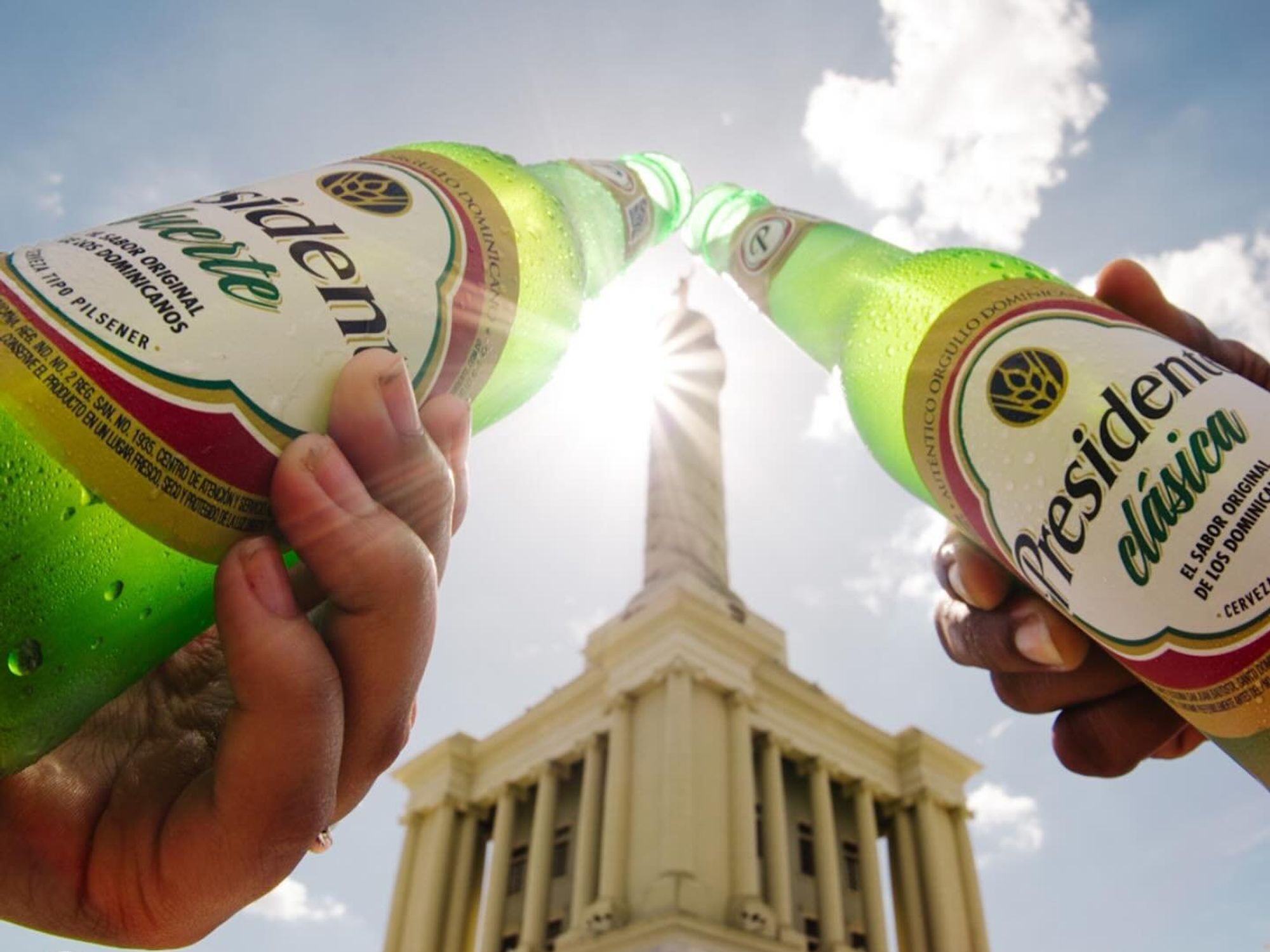 Two individuals raise bottles of 'Presidente,' Dominican Republic's beer in a toast.
