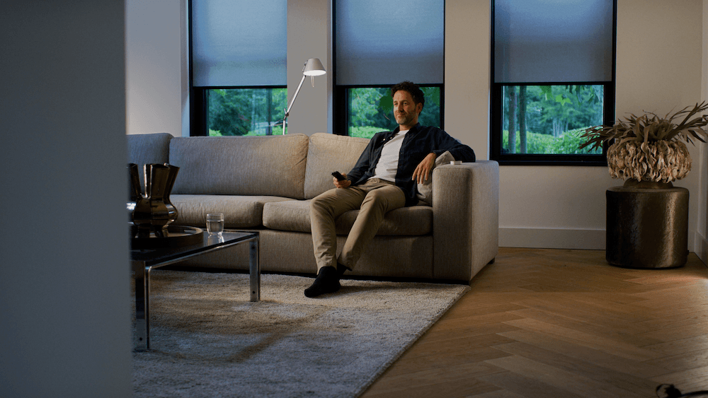 a photo of a man sitting on a couch in a room with Eve MotionBlinds on the windows
