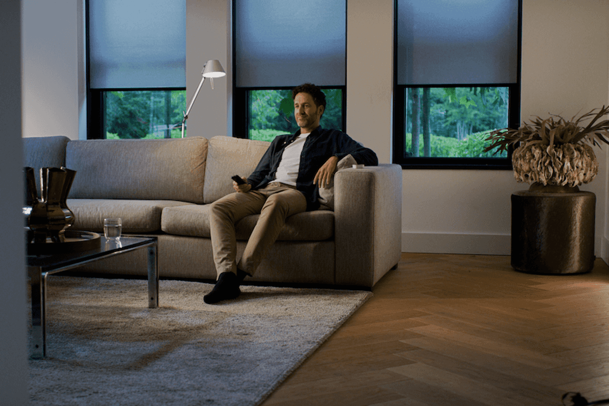 a photo of a man sitting on a couch in a room with Eve MotionBlinds on the windows