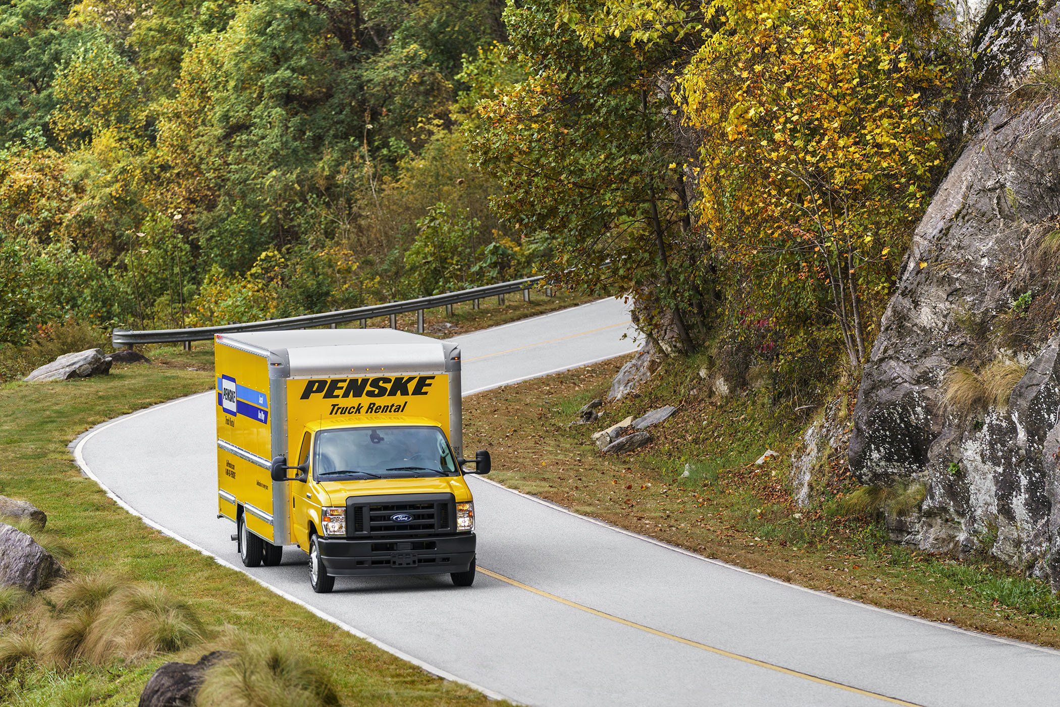 A yellow 16 foot Penske box truck drives along a curvy road surrounded by green trees.