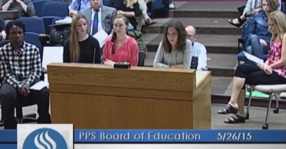Four teenagers stand at a wooden podium at a school board meeting