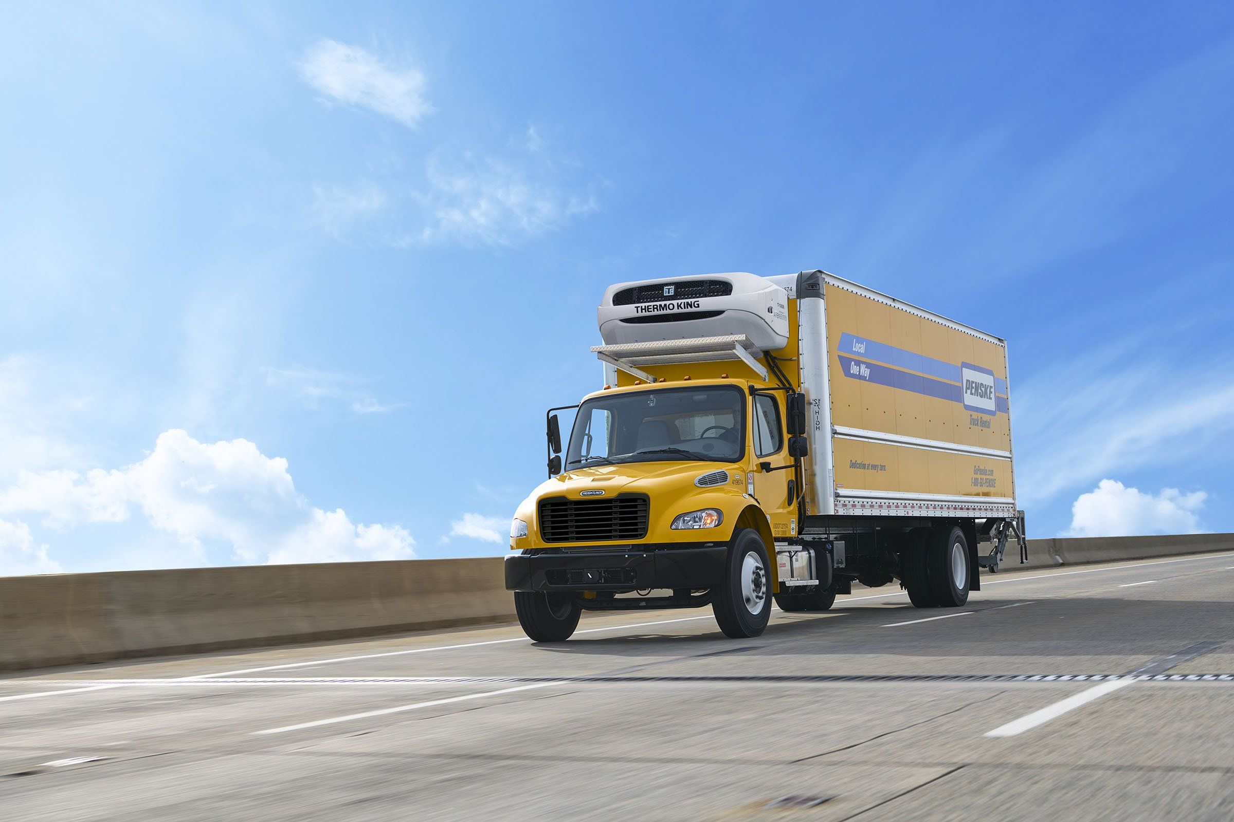Yellow Penske reefer truck driving over a cement bridge on a sunny day.