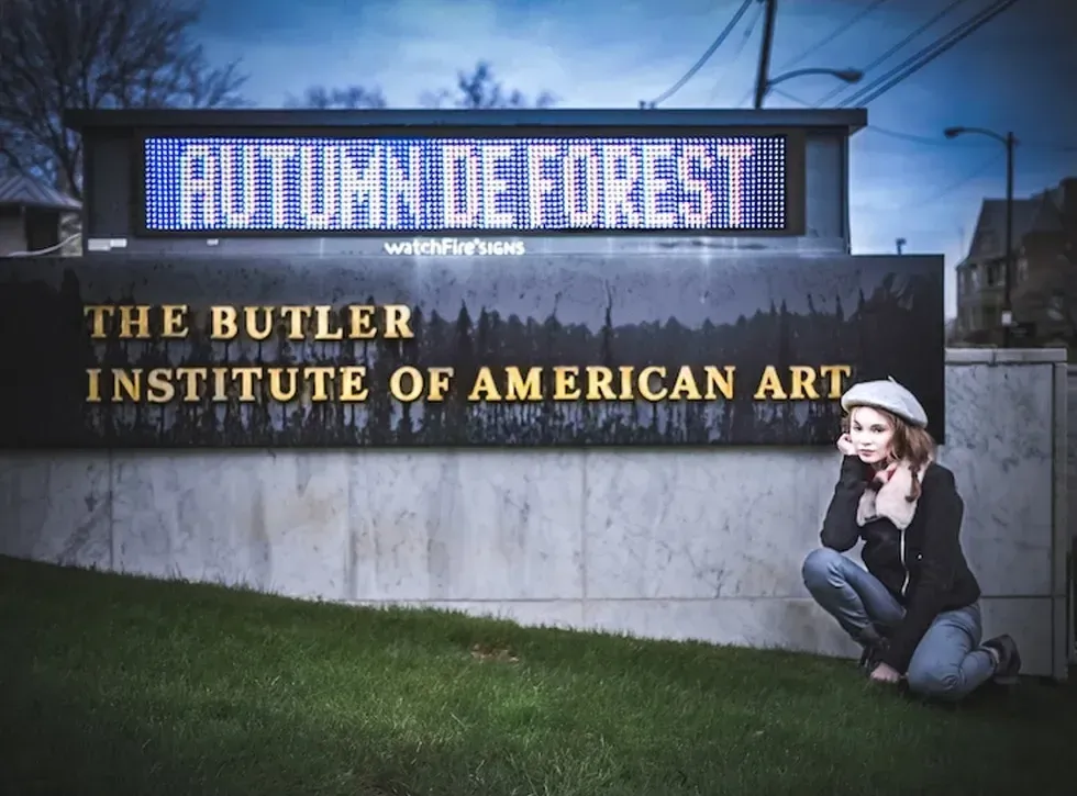 Woman kneeling beside Butler Institute sign with "AUTUMN DE FOREST" displayed above.