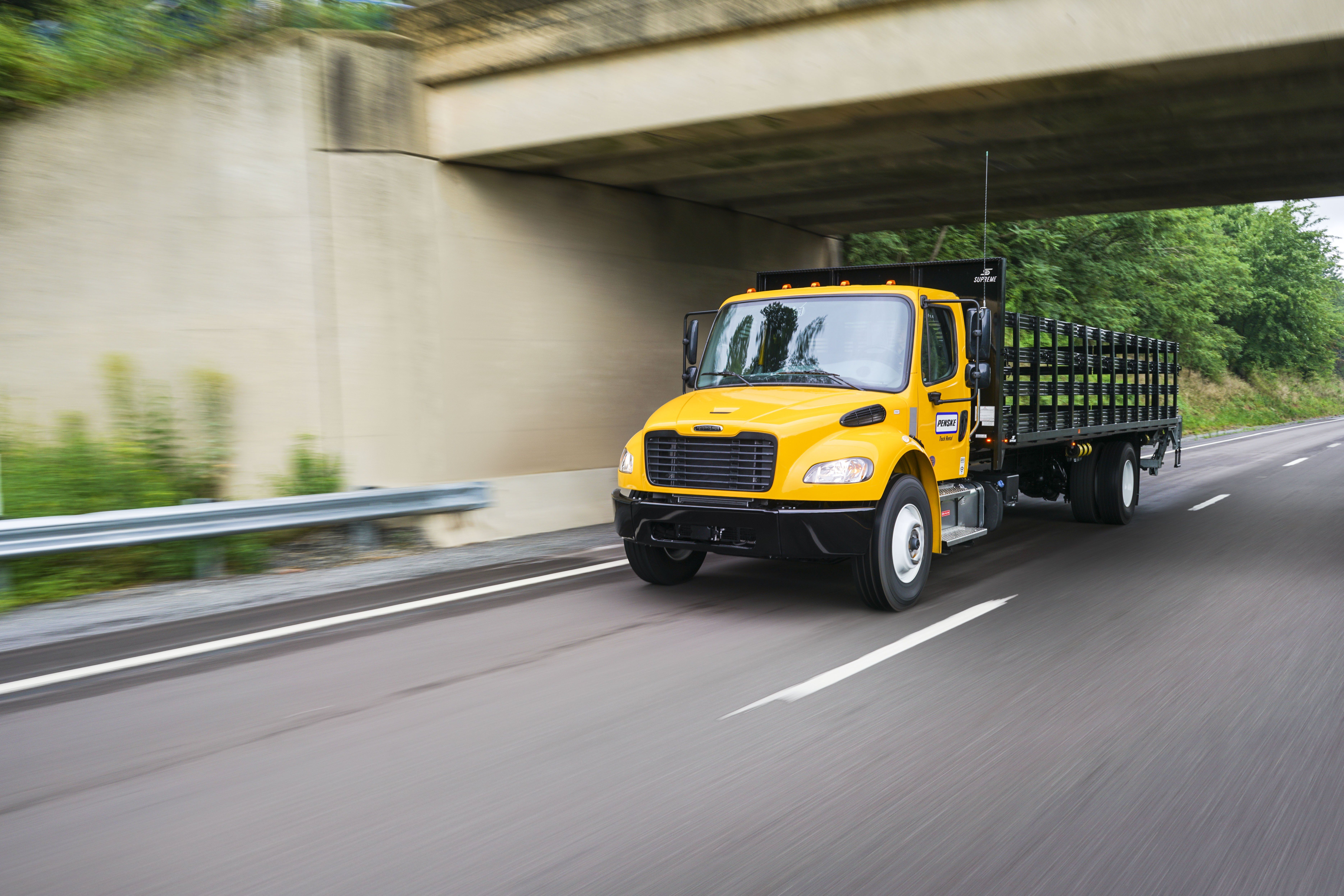 A yellow Penske flatbed truck drives on a freeway under a bridge.