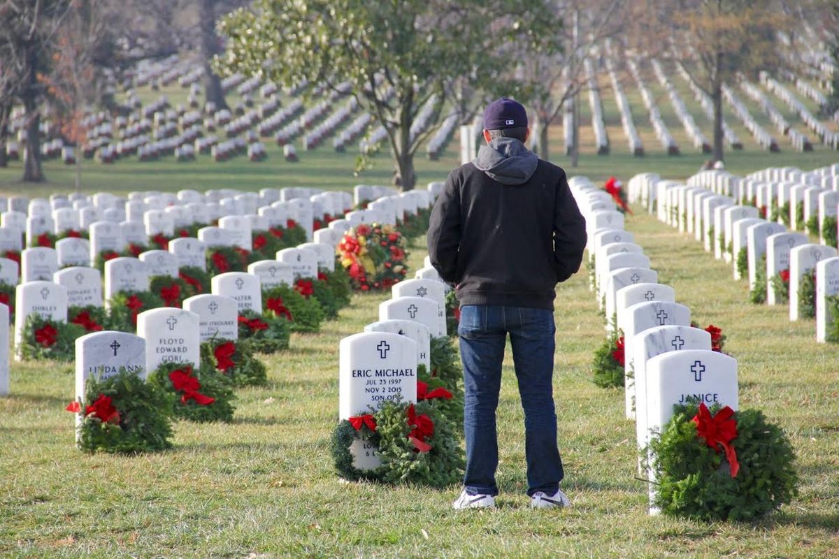 A person stands solemnly at a military cemetery surrounded by rows of wreath-adorned headstones, honoring veterans as part of Wreaths Across America.