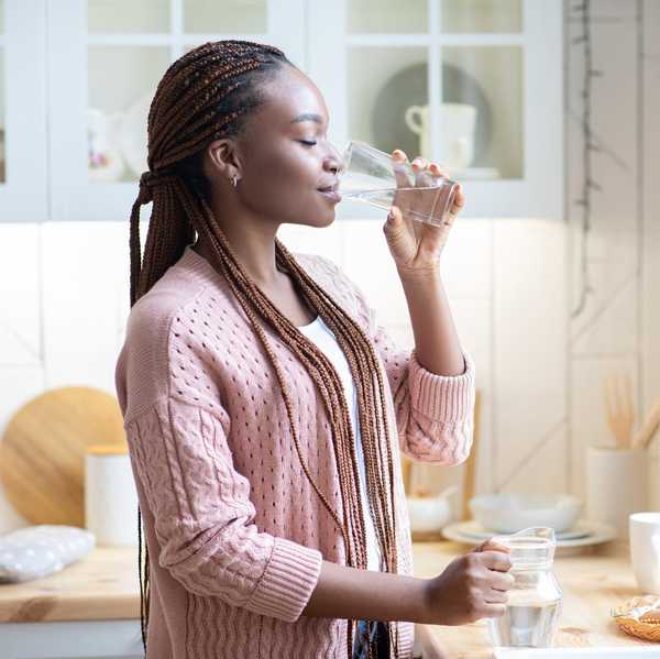 Thirsty African American Woman Drinking Water From Glass In Kitchen At Home, Young Black Female Standing Near Counter And Enjoying Heathy Refreshing Drink, Side View With Copy Space